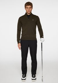 Tummanvihreä vetoketjullinen neuletakki, jossa on taitettu helma ja hihansuut, mustan kauluspuseron päällä, yhdistettynä mustiin housuihin ja golf-kengiin, pitäen kädessään golfmailaa.