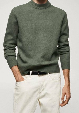 Mann trägt einen grünen Langarmpullover, der leicht in eine beige-weiße Jeans mit schwarzem Gürtel gesteckt ist, eine Hand in der Tasche.