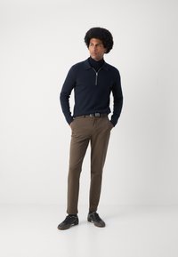 Marineblå glidelåsgenser, brune bukser, svarte sko. Glatt strikket tekstur, slim-fit design og kontrastbelte-detaljer.