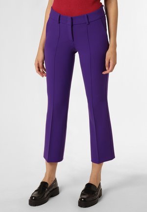 Žena nosící fialové kalhoty na míru s předními švy, v kombinaci s černými mokasínami a červeným topem, stojící na jednobarevném pozadí.