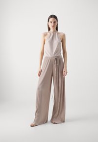 Light taupe pleated jumpsuit with halter neck, adjustable waist drawstring, and wide leg design, featuring a smooth, textured fabric.