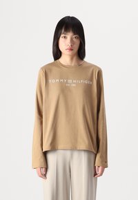 Braunes Langarm-T-Shirt mit weißem "TOMMY HILFIGER" Logo, Rundhalsausschnitt, lässiger Schnitt und gerippten Bündchen, kombiniert mit beigen Hosen.