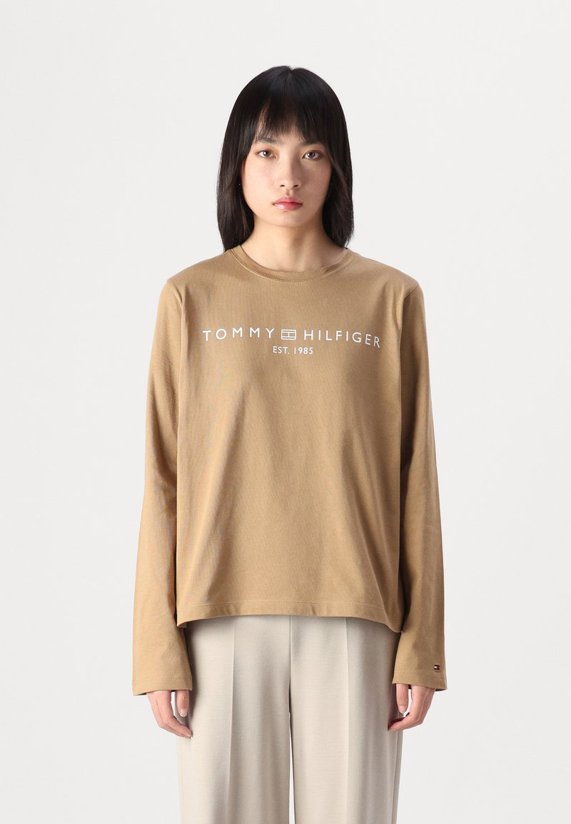 Braunes Langarm-T-Shirt mit weißem "TOMMY HILFIGER" Logo, Rundhalsausschnitt, lässiger Schnitt und gerippten Bündchen, kombiniert mit beigen Hosen.