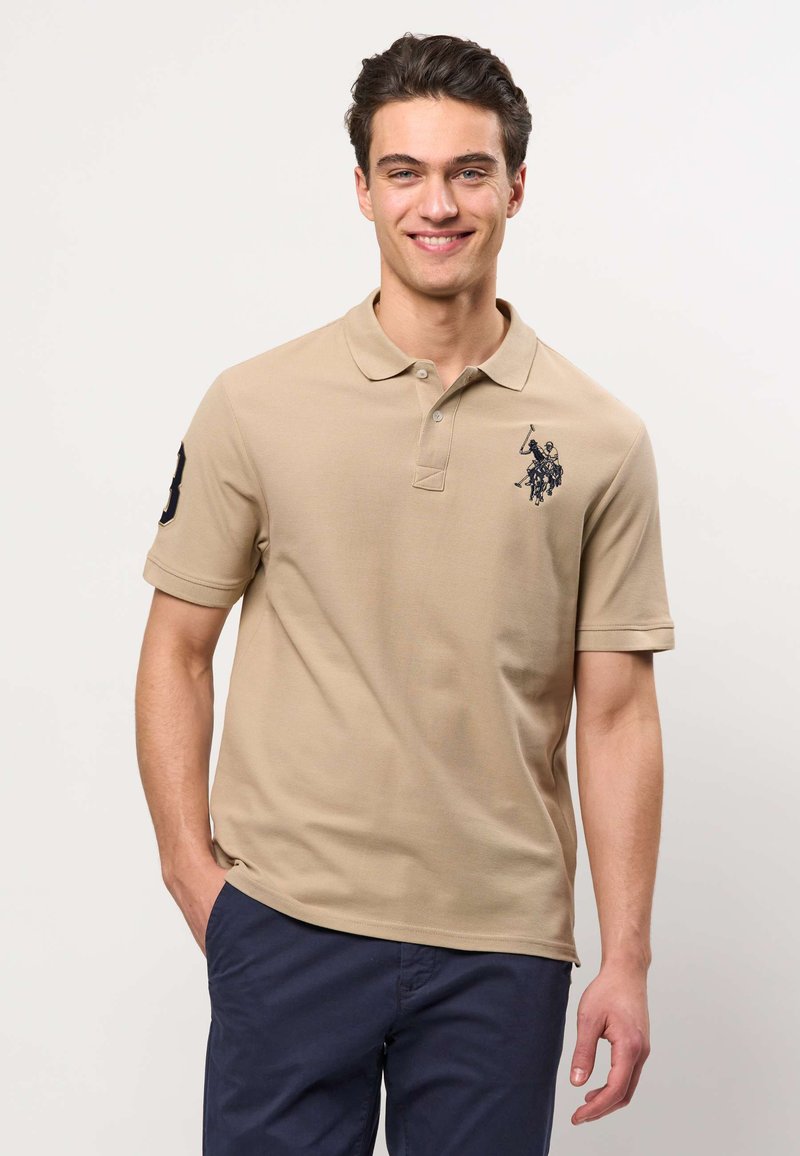 Jeune homme souriant, portant un polo beige avec un logo cheval et cavalier et un pantalon bleu marine, debout avec la main gauche dans la poche.