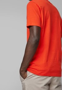 Koszulka z krótkim rękawem w jaskrawo pomarańczowym kolorze, wykonana z bawełny, z okrągłym dekoltem, gładką fakturą i luźnym krojem. Widok z boku pokazujący włożony do spodni krój.