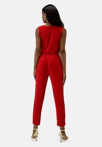 Mono rojo con cintura ajustada, diseño sin mangas y piernas estrechas. Tejido suave y largo hasta el tobillo, combinado con tacones altos beige.