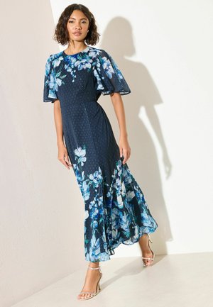 Nő tengerészkék virágos midi ruhát visel lebegő ujjakkal és bézs magassarkú szandállal, egyszerű fal előtt áll.