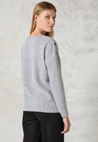 Grauer gestrickter Pullover mit lockerer Passform, Rundhalsausschnitt und Knopfdetails an der Schulter. Kombiniert mit schwarzen Hosen, die texturierte Stoffe sichtbar machen.