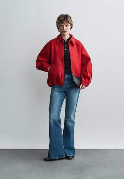 Άτομο που φοράει κόκκινο μπουφάν πάνω από μαύρο πουκάμισο, μπλε καμπάνα τζιν, μαύρα παπούτσια και κρατάει μια μικρή μαύρη τσάντα πάνω σε απλό φόντο.