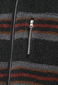 Nahaufnahme einer Fleecejacke mit horizontalen grauen, schwarzen, braunen und roten Streifen, ausgestattet mit einem vertikalen silbernen Reißverschluss und einer Reißverschlusstasche.