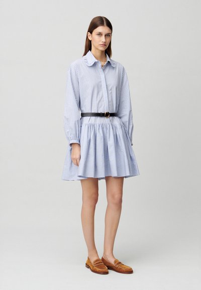 Abito-shirt a quadri blu chiaro con gonna arricciata, maniche lunghe e una cintura nera con fibbia rotonda. Mocassini marroni completano il look.
