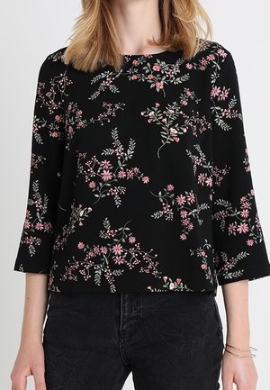 Kvinde iført sort top med pink og hvide blomsterprint og trekvartlange ærmer, kombineret med sorte bukser.