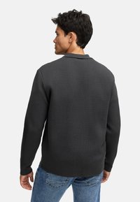 Man i en mörkgrå långärmad tröja och blå jeans, vänd bort, mot en enfärgad ljusgrå bakgrund.