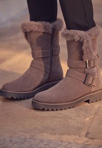 Bottines en daim gris avec bordure en fausse fourrure, fermeture éclair latérale et deux sangles horizontales ornées de boucles argentées. Semelle robuste pour une meilleure traction.