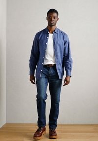 Μπλε πουκάμισο με κουμπιά και μακριά μανίκια, λευκή μπλούζα από κάτω, συνδυασμένο με σκούρα τζιν και καφέ μπότες με κορδόνια, σε στάση με φόντο ουδέτερης απόχρωσης.