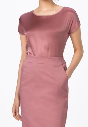 Femme portant un haut en satin rose poussiéreux à manches courtes rentré dans une jupe crayon taille haute assortie, une main dans la poche.