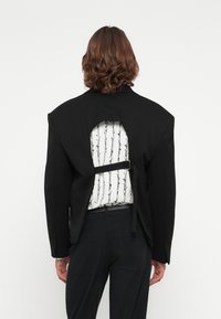 Schwarze Jacke mit einem ausgeschnittenen Rücken, der ein weißes Shirt mit einem schwarzen Stacheldraht-Muster zeigt, das mit einem Riemen gesichert ist.
