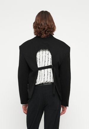 Chaqueta negra con un corte en la espalda que revela una camiseta blanca con un patrón de alambre de espino negro, asegurada con una correa.