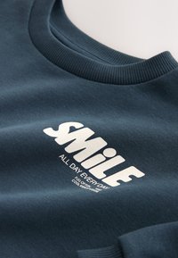 Sudadera azul oscuro con texto blanco "Smile all day every day full of fun cool kidsworks" en la parte delantera cerca del cuello.