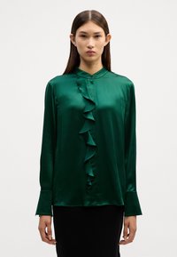Grüne Satinbluse mit Rüscheneinsatz an der Vorderseite, geknöpftem Kragen und langen Ärmeln. Glatte Textur und taillierte Passform mit einem dezenten Glanz.