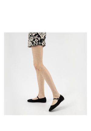 Nogi w czarnych balerinach Mary Jane z paskami przy kostce, w połączeniu z czarno-białą spódnicą w wzory, stojące na białym tle.