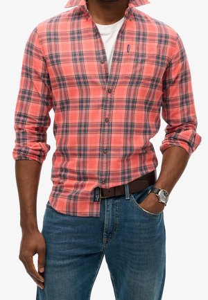 Hombre con camisa de cuadros rosa coral y azul marino con mangas remangadas, camiseta blanca debajo, vaqueros azules, cinturón marrón y reloj de pulsera plateado.