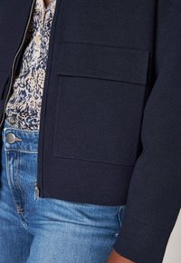 Giacca zip-up blu navy con grande tasca a pattina indossata sopra una camicia a fiori e jeans blu sul torso e braccio della persona.
