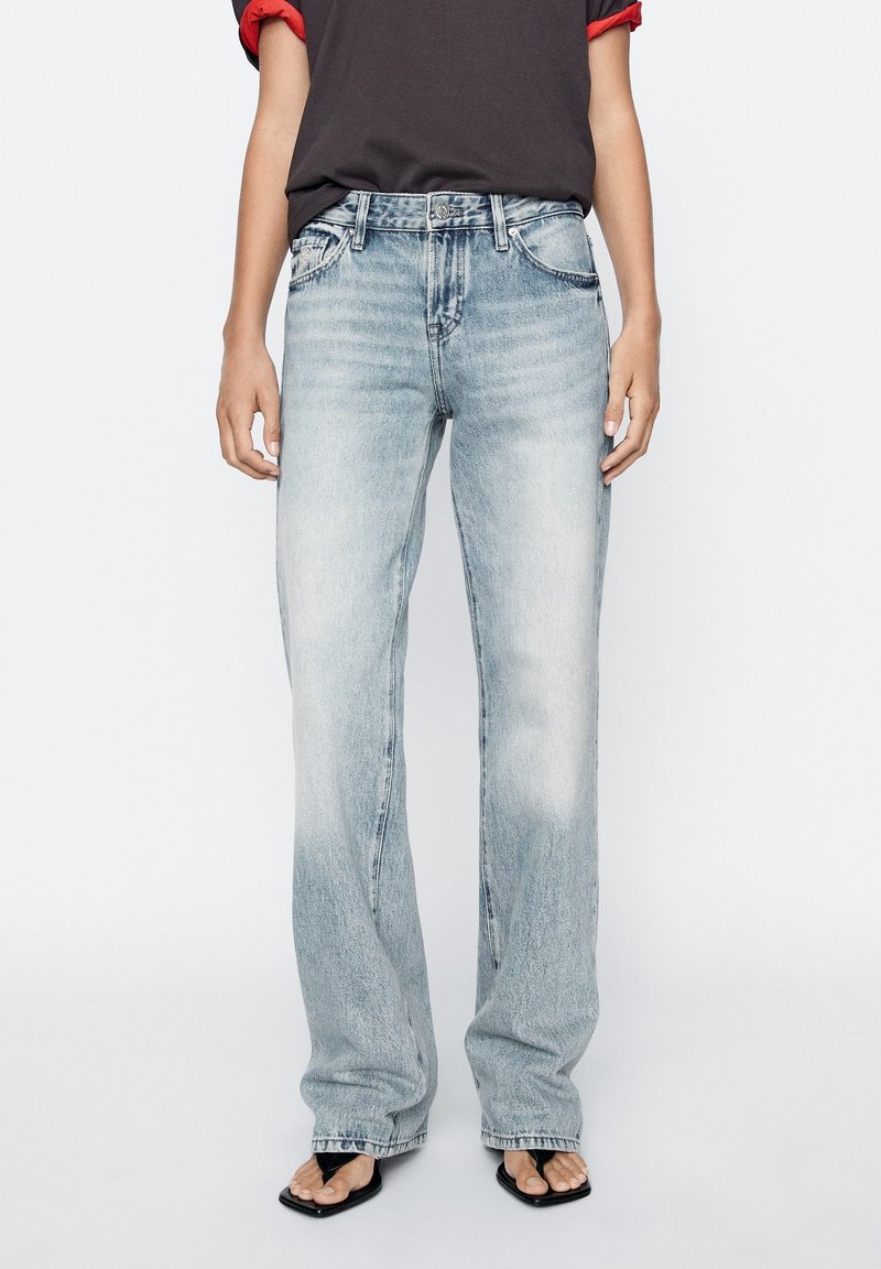 Personne portant un jean droit taille haute clair avec un t-shirt noir rentré et des sandales noires à bout carré.