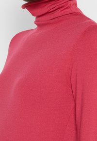 Rode coltrui van een zachte, rekbare stof. Heeft een getailleerd ontwerp en een geribbelde kraag. Naaddetails langs de zijkanten.