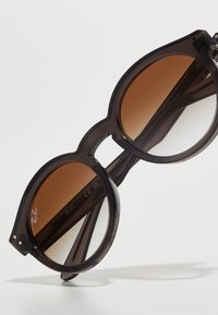Ray-Ban aurinkolasit, joissa on tummanharmaat läpikuultavat kehykset ja liukuväriset ruskeat linssit; pyöreä muoto ja hienovarainen logotieto linssissä.