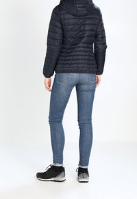 Svart puffjacka med horisontell sömnad och huva, ihop med blå skinny jeans och svarta träningsskor med grå detaljer.