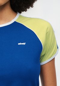 Το μπλε βαμβακερό μπλουζάκι διαθέτει μανίκια σε πράσινο και μπλε χρώμα, στρογγυλό λαιμό με γαλάζιο περίγραμμα και λευκό κεντημένο λογότυπο στο στήθος.