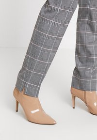 Szare spodnie w kratę o tapered momencie z rozcięciami przy kostkach, zestawione z eleganckimi, beżowymi, lakierowanymi szpilkami z wysmuklającym noskiem.