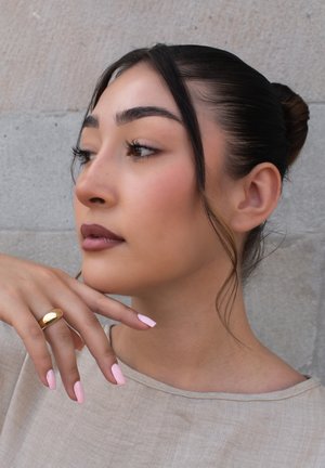 Nahaufnahme einer Frau mit langen braunen Haaren, die zu einem Dutt frisiert sind. Sie trägt ein beige Oberteil, hat hellrosa Nägel und einen goldenen Ring an ihrem Finger.