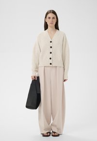 Frau trägt eine beige Strickjacke und Hosen mit weitem Bein, hält eine schwarze Tasche und steht vor einem schlichten weißen Hintergrund.