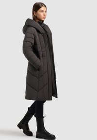 Mujer vistiendo un abrigo acolchado negro largo con capucha, leggings negros y botas negras hasta la mitad de la pantorrilla, de pie contra un fondo liso.