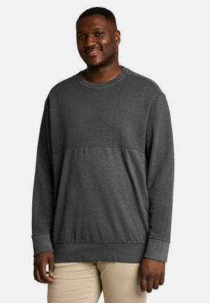 Mann lächelt, trägt ein dunkelgraues Sweatshirt mit langen Ärmeln und hellbeige Hosen vor einem einfarbigen hellen Hintergrund.