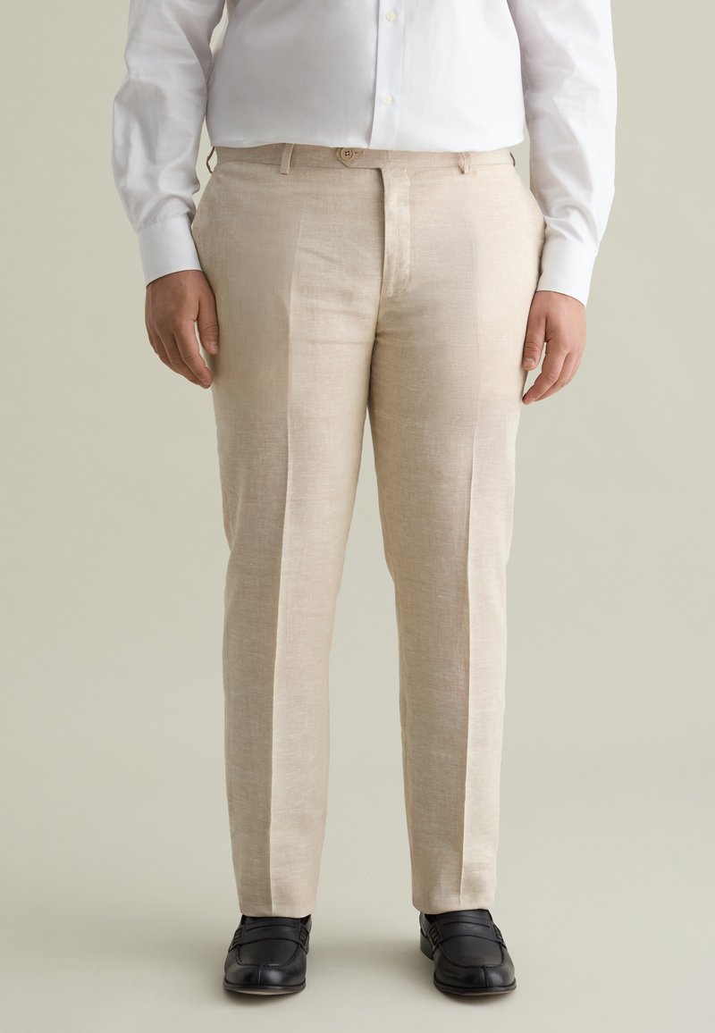 Beige linnenbroeken met een rechte voorkant, twee voorzakken en een subtiele textuur. Gecombineerd met een wit overhemd met lange mouwen en zwarte schoenen.