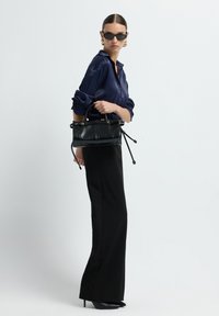 Femme en pantalon large noir et chemise en soie bleu marine, tenant un sac à main noir, portant des lunettes de soleil noires et des boucles d'oreilles en or, sur un fond uni.