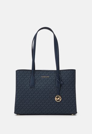Granatowa torba typu tote Michael Kors z podwójnymi paskami na ramię, powtarzający się wzór logo MK, złota zawieszka z logo i napisem „Michael Kors” na przodzie.