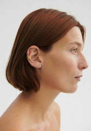 Rövid barna hajú nő arany virágos fülmandzsettát visel a bal fülén, oldalról látható egyszerű világos háttér előtt.