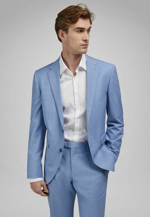 Junger Mann in einem hellblauen Anzug und weißem Hemd, der mit einer Hand in der Tasche steht und nach rechts blickt vor einem schlichten grauen Hintergrund.