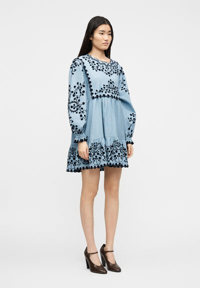 Γυναίκα που φοράει ανοιχτό μπλε φόρεμα με μαύρη κεντητή floral διακόσμηση, μακριά φουσκωτά μανίκια και σκούρα καφέ ψηλοτάκουνα παπούτσια Mary Jane, όρθια.