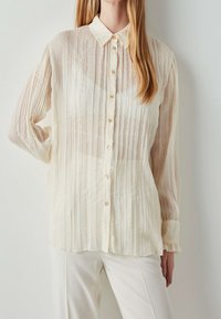 Chemise crème clair à boutons avec plis verticaux, texture douce et manches longues. Dotée d'un col et de boutons délicats en accents.