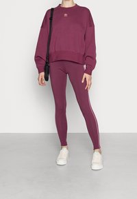 Žena oblečená v bordovém svetru Adidas a legínách s růžovými pruhy, v bílých teniskách a s černou křížovou taškou, stojící na šedé podlaze.