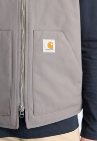 Szary kamizelka robocza z suwakiem z przodu, posiadająca dwie duże kieszenie oraz logo Carhartt w pomarańczowym na białym tle.