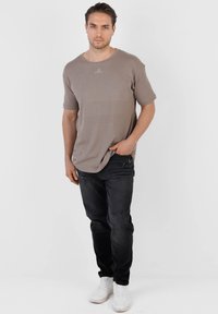 Jeune homme debout, main dans la poche, portant un t-shirt taupe, un jean noir et des baskets blanches, sur un fond clair uni.