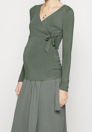 Femme enceinte portant un haut à manches longues style portefeuille vert olive avec un nœud noué sur le côté et une jupe assortie.