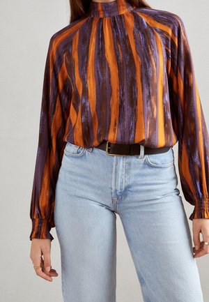 Kvinde iført en bluse med høj hals og lange ærmer med lodrette orange, lilla og brune striber, stoppet ned i lyseblå jeans med sort bælte.