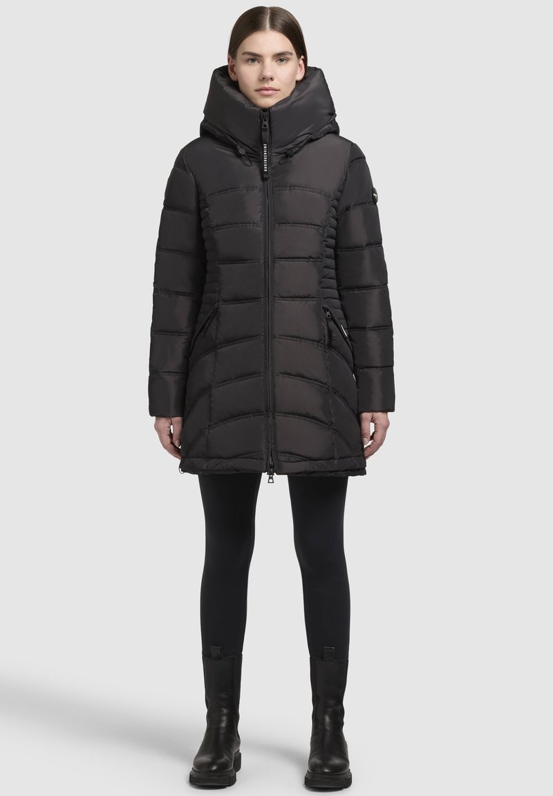 Doudoune noire avec un col haut et des détails de couture visibles, dotée d'une fermeture éclair sur le devant, de poches latérales et d'une coupe mi-genoux.